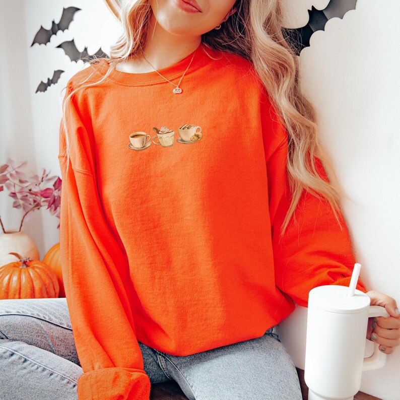 May include: Bright orange sweatshirt featuring a watercolor design of a teacup, teapot, and coffee cup. The sweatshirt is a solid color and has a crew neck. The model is wearing blue jeans and holding a white mug. Halloween decorations are in the background.