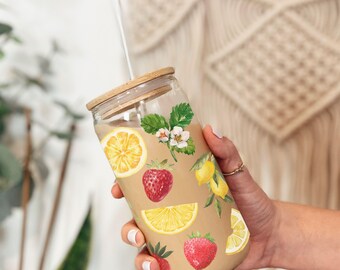 Op de afbeelding: Een herbruikbare glazen tumbler met een bamboe deksel en een rietje. De tumbler is versierd met een patroon van citroenen, aardbeien en bloemen.