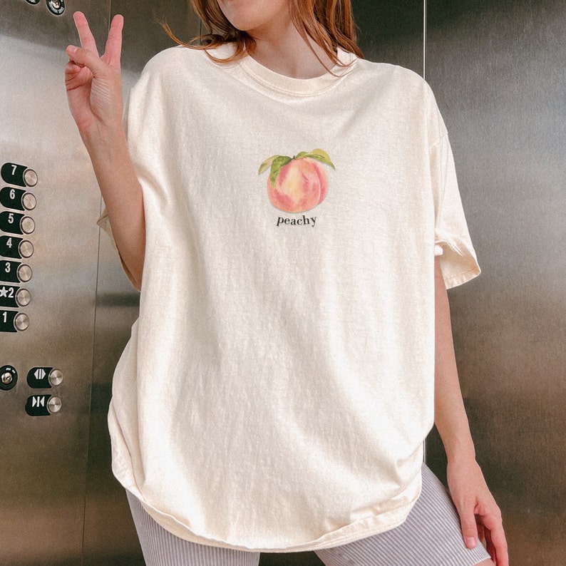 May include: A cream colored t-shirt with a peach graphic and the text "peachy" printed on the front.
