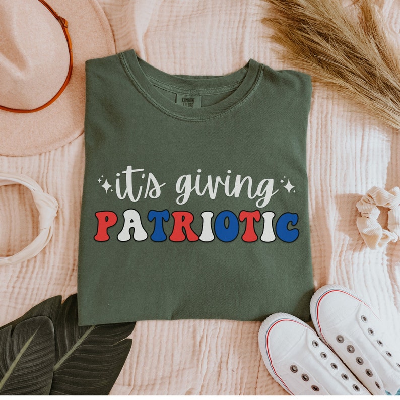 Pu&ograve; includere: Una maglietta verde oliva con la scritta "it's giving PATRIOTIC" in lettere bianche, rosse e blu. La maglietta &egrave; piegata e presentata con un cappello, un elastico per capelli e scarpe da ginnastica bianche.