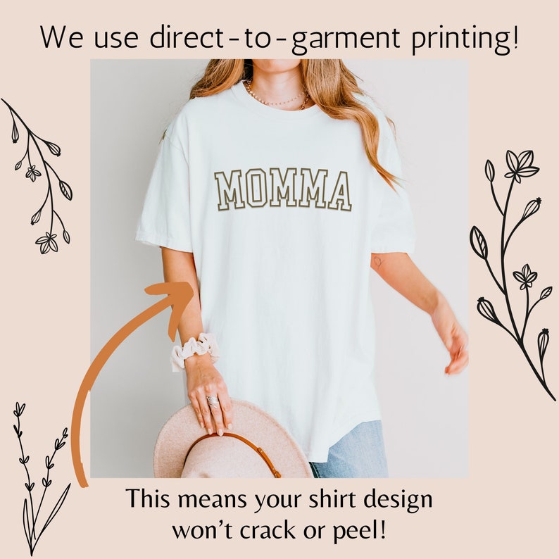 May include: White t-shirt with the word "MOMMA" printed in brown on the front. The text is outlined in black. The shirt is being worn by a person. The image includes text that says "We use direct-to-garment printing! This means your shirt design won't crack or peel!"