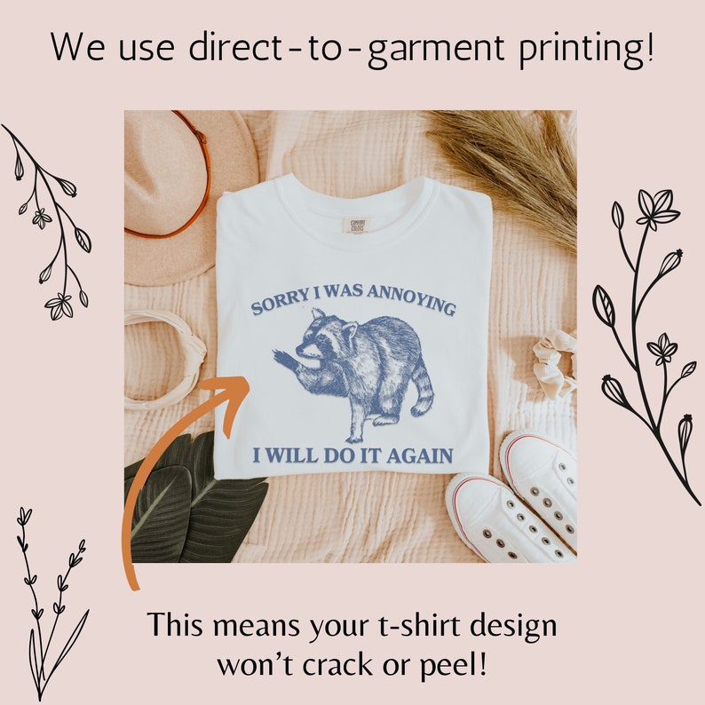 May include: White t-shirt with a blue raccoon graphic and the text "Sorry I was annoying, I will do it again". The t-shirt is on a light brown surface with a hat and a pair of white shoes.