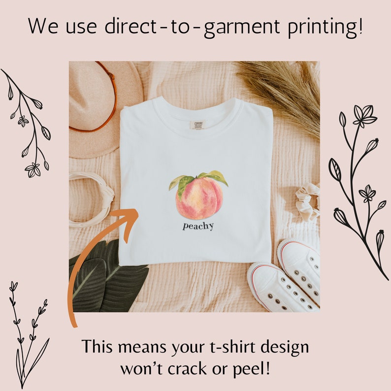 May include: White t-shirt with a peach illustration and the text "peachy" printed on the front. The t-shirt is laid out on a light brown surface with a hat and a pair of white shoes.
