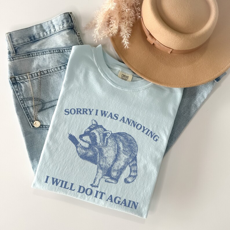 May include: A light blue t-shirt with a raccoon graphic and the text "Sorry I was annoying I will do it again".