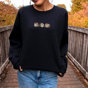 May include: Black crewneck sweatshirt featuring embroidered coffee cup designs. The embroidered cups include a coffee cup, a cappuccino, and a cup with whipped cream. The sweatshirt is worn by a person on a wooden bridge.