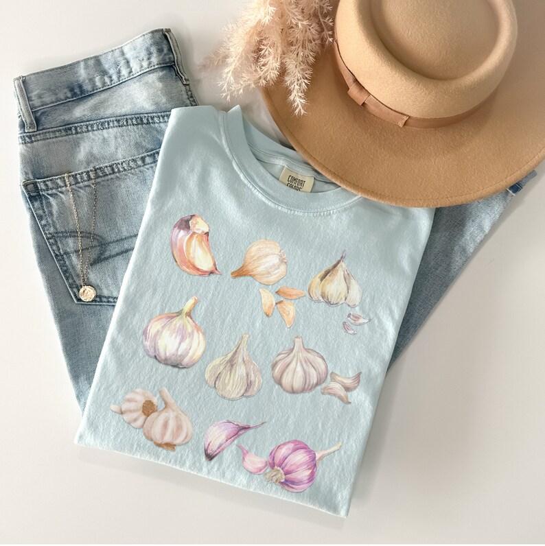 May include: A light blue t-shirt with a watercolor illustration of garlic cloves. The shirt has a small tag with the text "Comfort Colors" on the front.