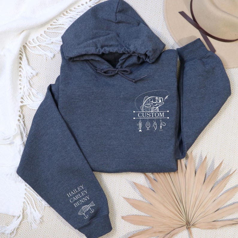 Pode incluir: Uma camisola de capuz cinzenta escura com um gr&aacute;fico branco de um peixe e iscas de pesca. O texto "CUSTOM" est&aacute; impresso abaixo do peixe. A manga tem os nomes "HAILEY, CARLEY, BENNY" impressos nela.