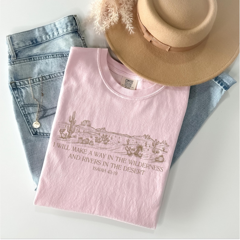 Puede incluir: Camiseta rosa claro con un gr&aacute;fico de paisaje des&eacute;rtico y el texto "I WILL MAKE A WAY IN THE WILDERNESS AND RIVERS IN THE DESERT ISAIAH 43:19". La camiseta se combina con vaqueros azules claros y un sombrero beige.