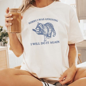 May include: White t-shirt with a blue illustration of a raccoon and the text "Sorry I was annoying I will do it again".