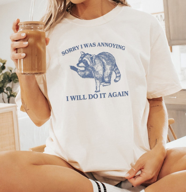 May include: White t-shirt with a blue illustration of a raccoon and the text "Sorry I was annoying I will do it again".