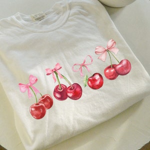 Puede incluir: Camiseta blanca con cinco lazos rosas y cerezas rojas. Cada cereza tiene un tallo y una hoja verdes.