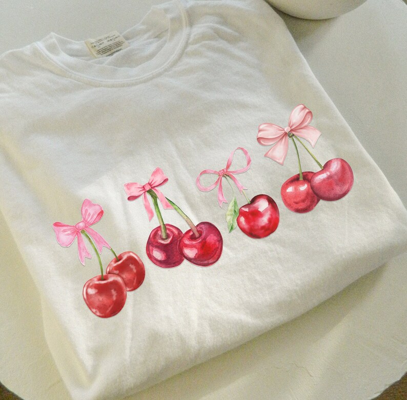 Puede incluir: Camiseta blanca con cinco lazos rosas y cerezas rojas. Cada cereza tiene un tallo y una hoja verdes.