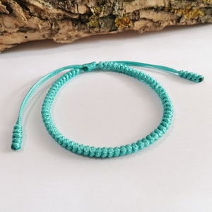May include: A turquoise blue braided bracelet with a knotted design. The bracelet is made of a thin cord and has a simple, elegant style.
