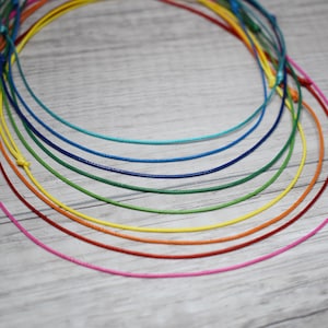 Puede incluir: Una colección de cordones finos y coloridos en un arcoíris de tonos, incluyendo rosa, rojo, naranja, amarillo, verde, azul y turquesa. Los cordones están dispuestos en un patrón circular sobre un fondo claro de vetas de madera.