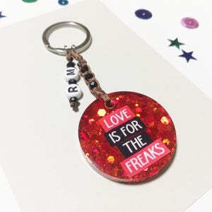 May include: Red glitter keychain with a silver key ring and brown string with white beads that spell "ROM". The keychain has a red glitter background with a white and black graphic that says "Love is for the Freaks".