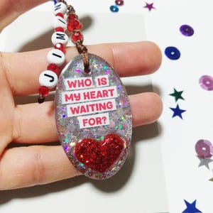 May include: A silver oval keychain with glitter and a red heart. The keychain has the text "Who is my heart waiting for?" on it. The keychain is attached to a string of red and white beads that spell out "Jimin".