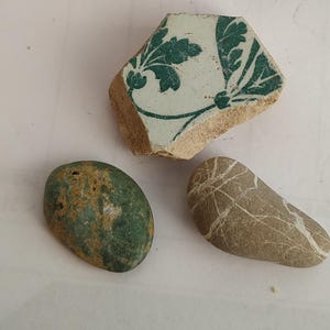 Può includere: Una collezione di tre oggetti composta da un frammento di ceramica bianca con un motivo floreale verde, una pietra verde liscia di forma ovale con una consistenza marrone maculata e una pietra color marrone chiaro con sottili linee bianche.