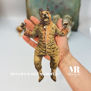 Puede incluir: Un títere de papel de un perro que lleva un uniforme militar verde y marrón con botones dorados. El títere está sostenido en la mano de una persona. El texto "DIY GIFT & SELF MADE FUN" es visible en la parte inferior de la imagen.