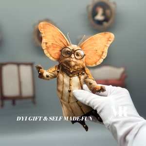 Peut inclure: Un modèle en papier d'une créature fantaisiste aux grandes oreilles, portant des lunettes et un manteau blanc. Le modèle est tenu dans une main gantée. Le texte "DIY GIFT & SELF MADE FUN" est visible sous le modèle.
