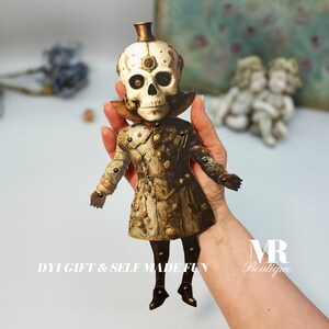 May include: A flat, articulated paper doll featuring a skull head with a small top hat and a detailed, ornate coat in shades of cream, brown, and gold. The figure has jointed limbs connected by small metal fasteners and includes the text DIY GIFT & SELF MADE FUN.