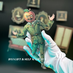 May include: A paper doll with a green suit and an apple head with a smiling face. The doll is being held by a gloved hand. The text "DIY GIFT & SELF MADE FUN" is visible at the bottom of the image.