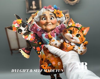 May include: A set of three articulated paper figures featuring an elderly person with white hair and a floral crown, a black llama with a floral wreath, and an orange tabby cat with a floral crown, all connected by metal brads. Text reads DYI GIFT & SELF MADE FUN.