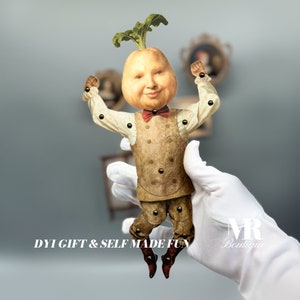 Puede incluir: Una peculiar figura de papel con cabeza de nabo, que lleva chaleco, pantalón y pajarita. La figura tiene articulaciones y está sujeta por una mano enguantada. El texto "DYI GIFT & SELF MADE FUN" es visible.