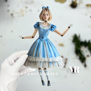 May include: A blue and white paper doll of Alice in Wonderland with a white lace trim on her dress. The doll has movable arms and legs. The doll is holding a white glove in one hand. The doll is standing on a white surface. The text "DIY GIFT & SELFMADE FUN" is visible at the bottom of the image.