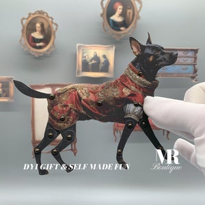 Puede incluir: Un perro de papel negro y marrón que lleva un abrigo estampado en rojo y dorado. El perro está en posición de pie con las patas dobladas a la altura de las rodillas. El perro es una marioneta con articulaciones móviles. El perro está delante de un fondo blanco con una mesa pequeña y cuadros enmarcados en el fondo. El texto "DIY GIFT & SELF MADE FUN" está en la parte inferior de la imagen. El texto "MR Boutique" está en la esquina inferior derecha de la imagen.