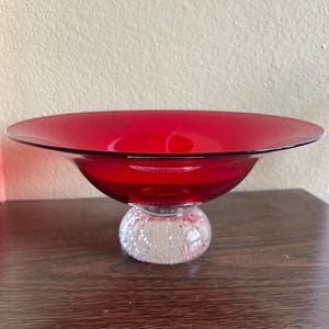 May include: A vibrant red glass bowl with a broad, shallow rim. It rests on a clear, spherical base filled with iridescent glitter. The bowl is likely for decorative use or serving. The bowl's diameter is approximately 25 cm.