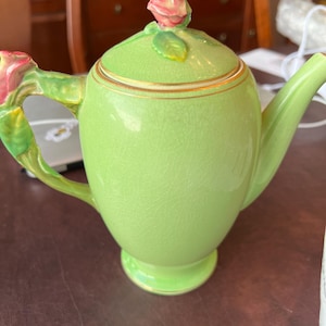 Puede incluir: Una tetera de cerámica verde con un adorno de capullo de rosa y ribete dorado. La tetera tiene un delicado diseño floral en el asa.