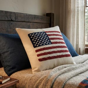 Puede incluir: Un cojín decorativo con un diseño de bandera estadounidense texturizado en rojo, blanco y azul. El cojín rectangular descansa sobre una cama con otras almohadas y una manta. La cama tiene un cabecero de madera.