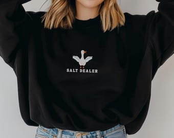 Puede incluir: Una sudadera negra con un gráfico de un ganso blanco y el texto "SALT DEALER" bordado en la parte delantera. La modelo lleva pantalones vaqueros azules.