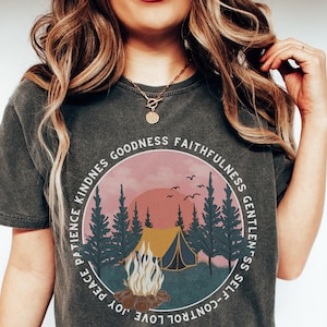 May include: A dark gray t-shirt with a circular graphic of a campfire scene in a forest. The graphic includes a tent, trees, a sunset, and birds. The words "Self-Control, Love, Joy, Peace, Patience, Kindness, Goodness, Faithfulness, Gentleness" are written around the graphic.