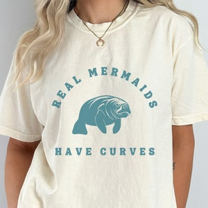 May include: A white t-shirt with a teal blue manatee graphic and the text "REAL MERMAIDS HAVE CURVES".