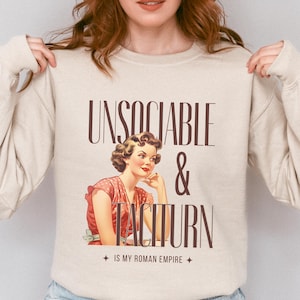 Puede incluir: Una sudadera beige con un diseño gráfico de estilo vintage. El gráfico presenta a una mujer con cabello rojo y un vestido de lunares rojos y blancos. El texto en el gráfico dice "Unsociable & Taciturn" y "Is My Roman Empire".