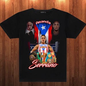 May include: Black t-shirt featuring a graphic of a female boxer with the Puerto Rican flag and the name "Amanda Serrano." The design includes images of the boxer and championship belts.