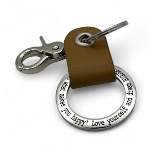 May include: A keychain featuring a tan leather strap attached to a silver metal ring engraved with the text: Do what makes you happy! Love yourself and dream BIGGER. The keychain includes a silver metal lobster clasp and a split ring attachment.