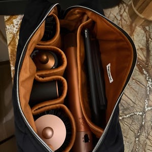 May include: A black zippered travel case with a tan interior, featuring multiple padded compartments. Inside are several hair styling tools, including a copper-colored hair dryer, a black flat iron with the word NOIRI, and various round brush attachments.