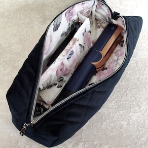 Pode incluir: Uma mala de viagem preta acolchoada com fecho de correr prateado. O interior é forrado com um tecido branco com um padrão floral rosa e cinzento. No interior, uma ferramenta de modelar o cabelo azul-escura e cobre está guardada junto a uma bolsa com a etiqueta NOIRI.