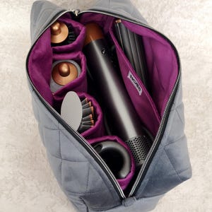 May include: A quilted gray storage bag with a vibrant purple interior lining and a silver zipper closure. The bag contains a hair styling tool with copper-colored attachments and a white fabric label with the text NOIRI stitched inside.