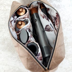 May include: A quilted beige velvet travel case with a zipper closure, containing a gray hair styling tool and several attachments. The interior is lined with a white fabric featuring a floral pattern and a small white tag labeled with the text NOIR.