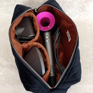 Puede incluir: Un bolso de almacenamiento acolchado negro con cremallera, forro interior color óxido y una etiqueta blanca con la palabra NOIR. En su interior, contiene un secador de pelo gris oscuro con cabezal circular rosa y dos accesorios grises.