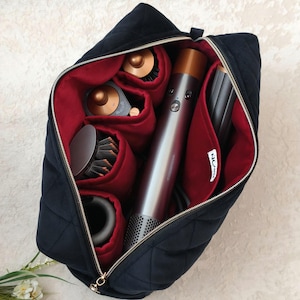 Peut inclure: Un sac de rangement noir matelassé avec une doublure intérieure rouge profond, conçu pour contenir un outil de coiffure et ses divers accessoires. Le sac présente une fermeture éclair dorée et une petite étiquette blanche avec le texte "NOM".
