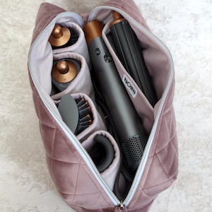 May include: A quilted mauve velvet storage bag with a white zipper, containing a gray hair styling tool with copper accents and several cylindrical attachments. A small white label with the black text NOIRI is sewn onto the interior divider.
