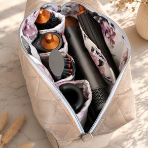 May include: A beige quilted travel organizer bag with a floral interior lining, containing a hair dryer, multiple styling attachments, and a brush. The interior fabric features a white background with purple and pink floral patterns and a small tag reading NOIR.