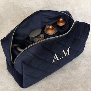 May include: A navy blue quilted velvet travel case with a silver zipper closure and the embroidered initials A.M. on the front. The open interior reveals multiple compartments containing various hair styling tool attachments in copper and gray.
