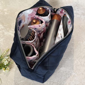 May include: A navy blue quilted cosmetic bag with a floral interior, open to reveal hair styling tools. The bag has a zip closure and is designed to hold various hair care accessories, such as brushes and a hair dryer.