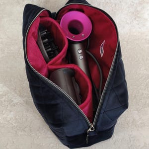 May include: A navy blue quilted velvet storage bag with a vibrant magenta interior lining, containing a grey hair dryer with a magenta circular head and two hair dryer attachments, secured by a silver metal zip closure with a small white tag.
