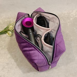 May include: A quilted, purple velvet travel bag with a silver zip, open to show a hair dryer and other styling tools. The bag has a diamond pattern and a light-coloured interior.